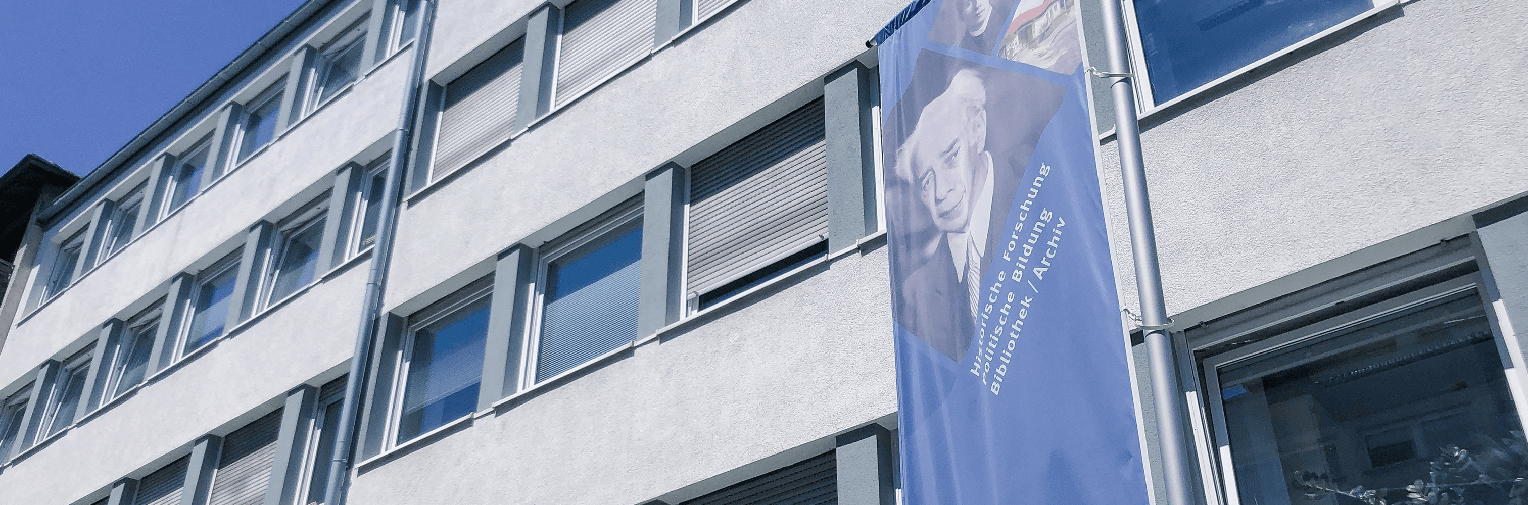 Außenfassade mit vielen Fenstern. Davor weht eine große Flagge mit Heuss und Informationen.