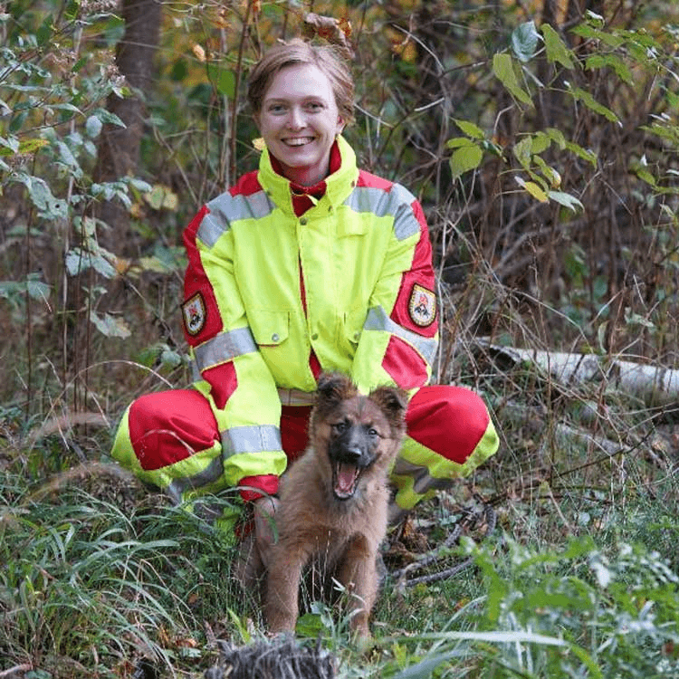 Porträtfoto einer Frau in gelb-roter Einsatzkleidung mit Reflektorstreifen und Abzeichen, vermutlich einer Rettungs- oder Hilfsorganisation. Sie kauert im Freien in einem Waldstück und lächelt in die Kamera. Vor ihr steht ein junger brauner Schäferhundwelpe.