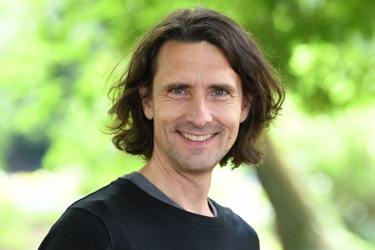 Porträtfoto eines Mannes mit schulterlangen braunen Haaren und blauen Augen, gekleidet in ein schwarzes T-Shirt. Er lächelt freundlich in die Kamera. Der Hintergrund zeigt unscharfes grünes Laub im Freien.