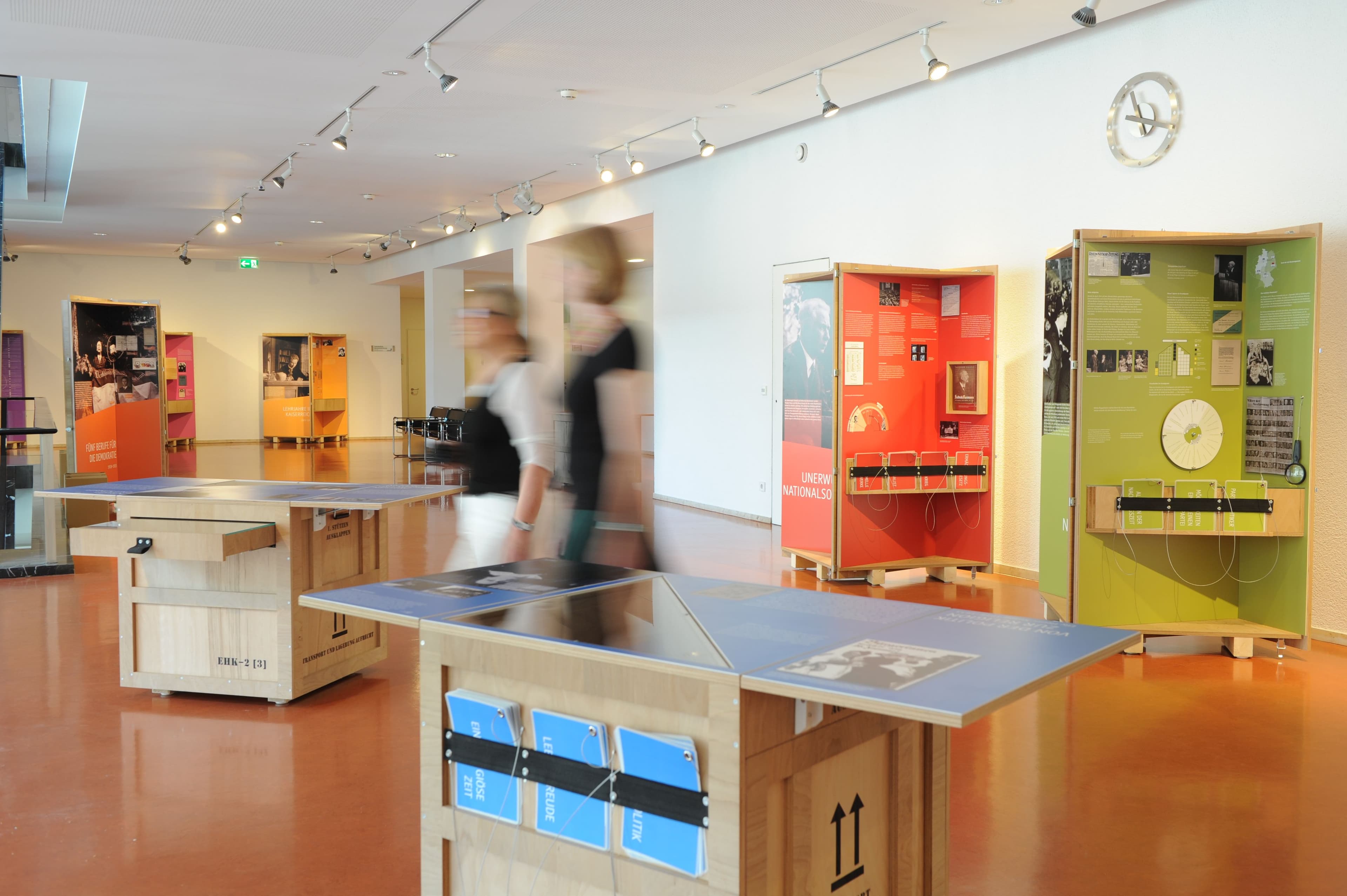 Zwei Besucherinnen gehen durch die Ausstellung Demokratie als Lebensform mit aufklappbaren Displays aus Holz.