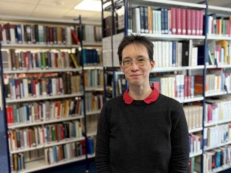 Porträtfoto einer Person mit kurzem dunklem Haar und einer Brille, gekleidet in einen schwarzen Pullover mit rotem Kragen. Sie lächelt leicht in die Kamera. Im Hintergrund sind mehrere Bücherregale mit zahlreichen Büchern in einer Bibliothek zu sehen.