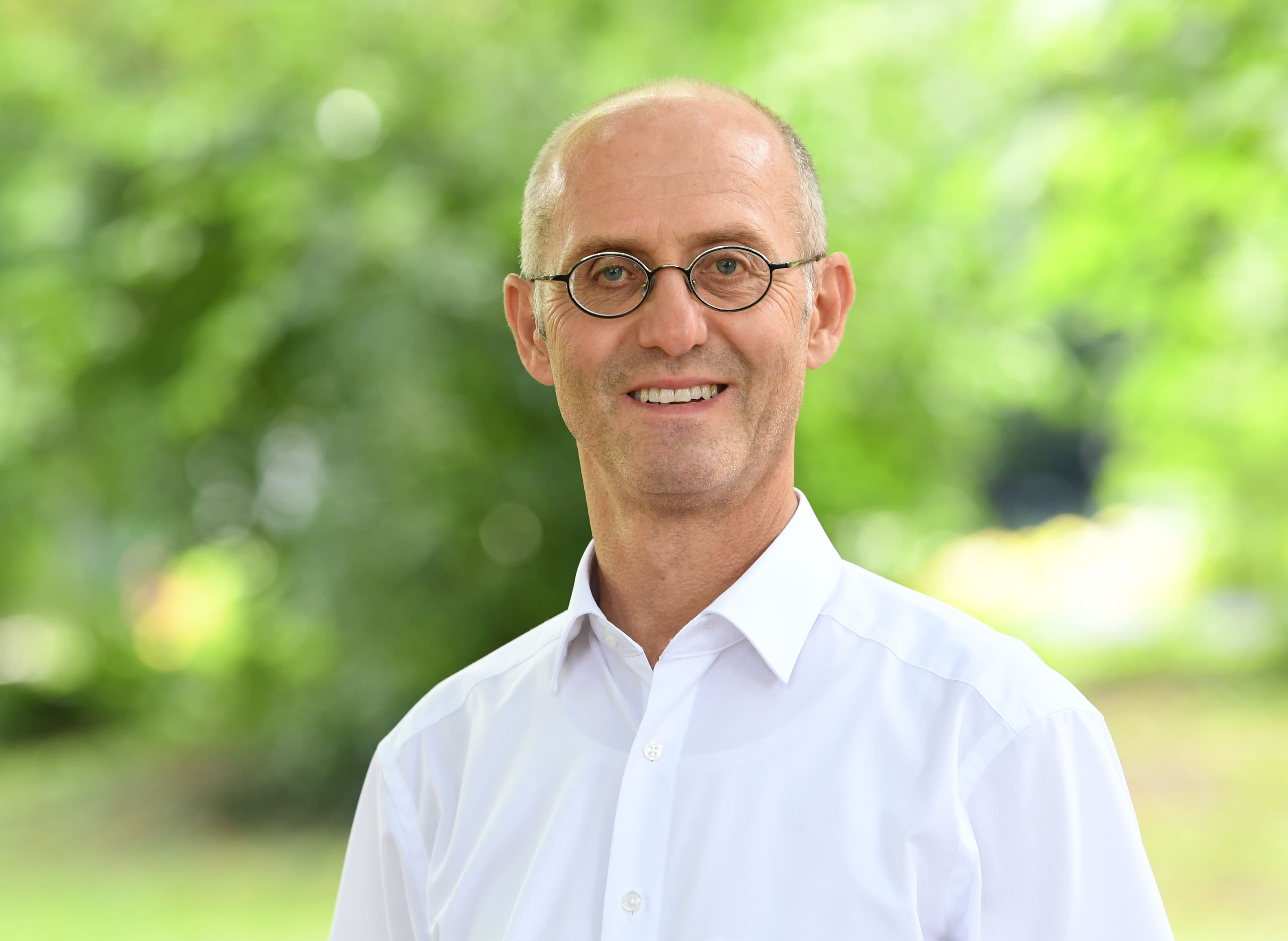 Porträtfoto von Dr. Ernst Wolfgang Becker mit kurzen grauen Haaren und einer randlosen Brille, gekleidet in ein weißes Hemd. Er lächelt freundlich in die Kamera. Der Hintergrund zeigt unscharfes grünes Laub im Freien.