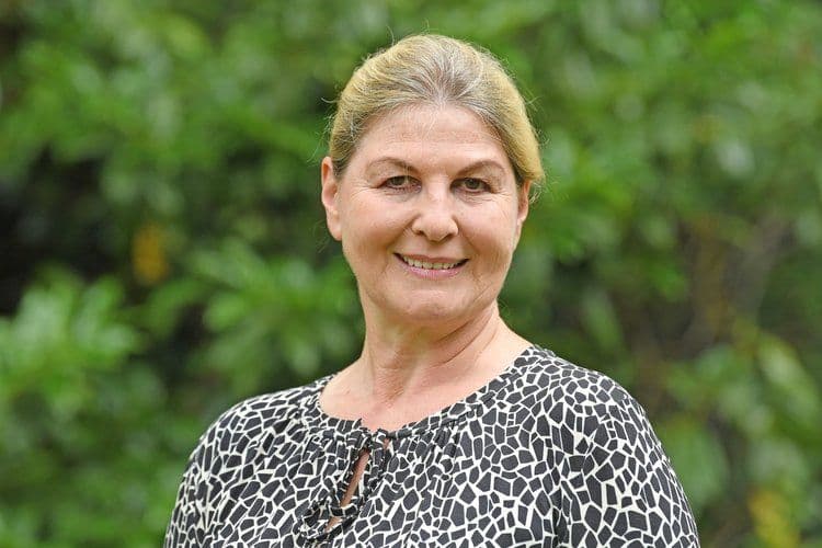 Porträtfoto einer Frau mit nach hinten gestecktem grau-blondem Haar, gekleidet in eine schwarz-weiß gemusterte Bluse. Sie lächelt freundlich in die Kamera. Der Hintergrund zeigt unscharfes grünes Laub im Freien.