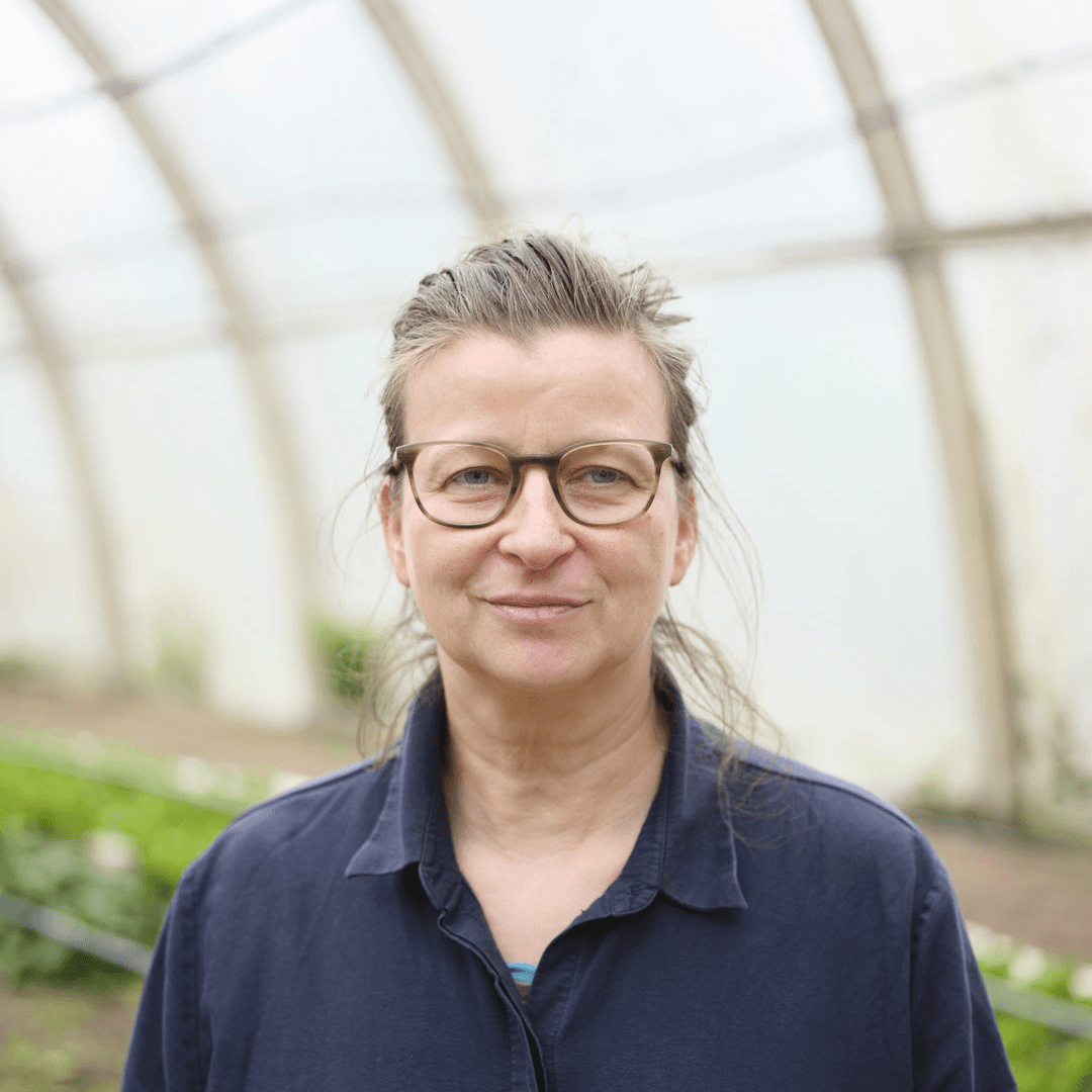 Porträtfoto einer Frau mit grau-braunem Haar und einer olivfarbenen Brille, gekleidet in ein dunkelblaues Hemd. Sie lächelt leicht in die Kamera. Der Hintergrund zeigt das Innere eines Gewächshauses mit Pflanzenreihen.