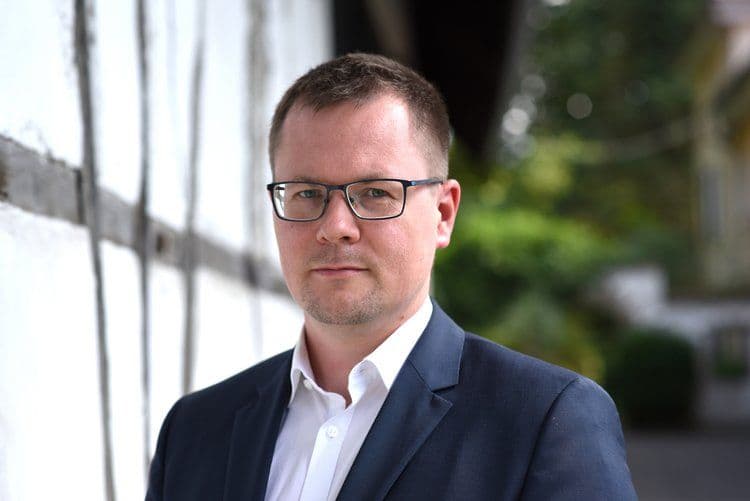 Porträtfoto von Dr. Jan Ruhkopf mit kurzem hellbraunem Haar und einer eckigen Brille, gekleidet in einen dunkelblauen Blazer und weißes Hemd. Er blickt ruhig in die Kamera. Der Hintergrund zeigt eine helle Wand und unscharfes Grün im Freien.