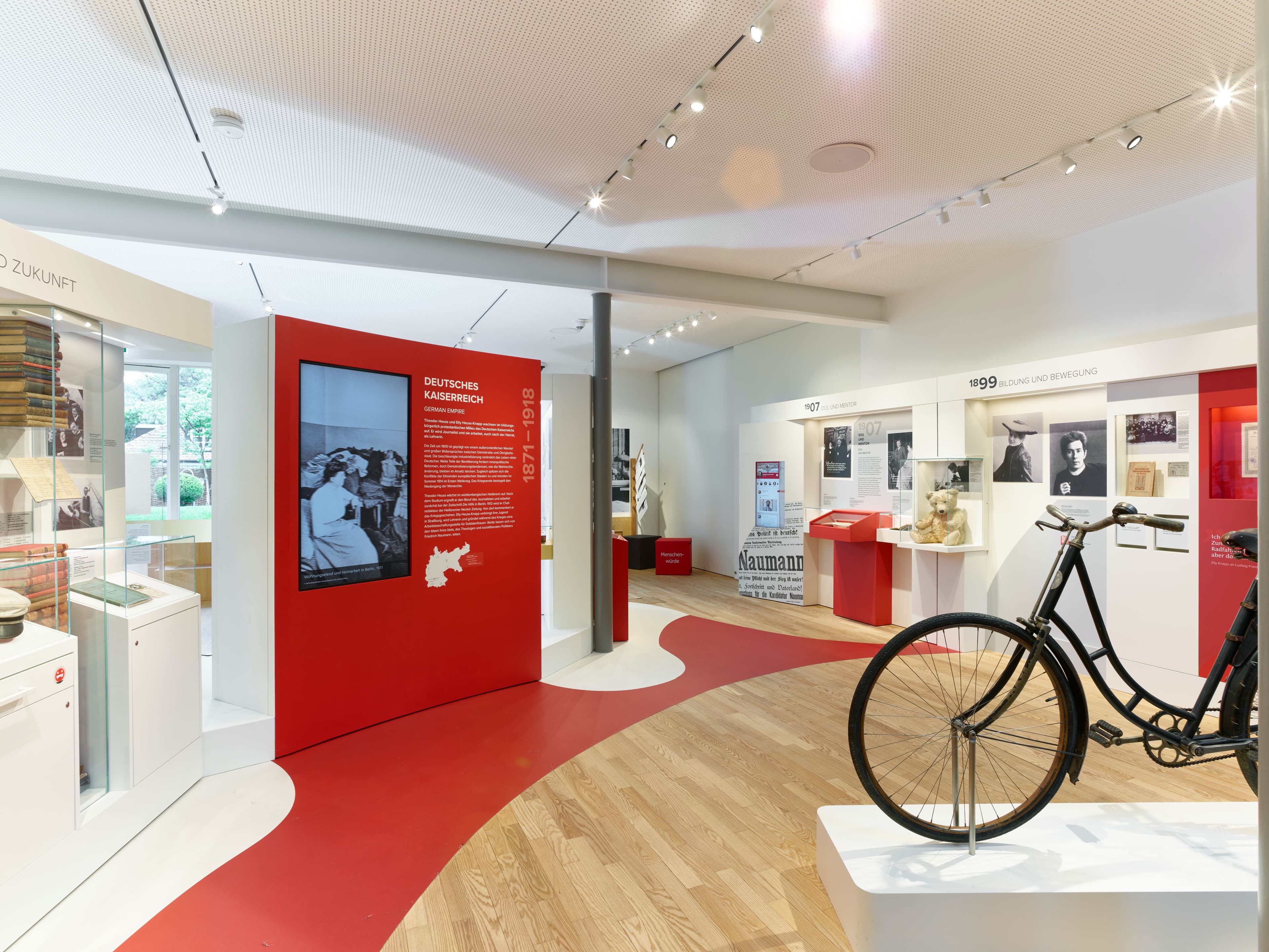 Innenansicht eines modernen Museums mit rotem Laufband auf hellem Holzboden. Im Vordergrund steht ein historisches Fahrrad auf einem weißen Sockel. In der Mitte dominiert eine rote Ausstellungswand zum Thema „Deutsches Kaiserreich 1871–1918" mit historischen Fotografien und Texten. Im Hintergrund sind weitere Ausstellungsbereiche mit Vitrinen, Texttafeln und Objekten aus dem späten 19. Jahrhundert sowie ein Teddybär als Exponat zu sehen.