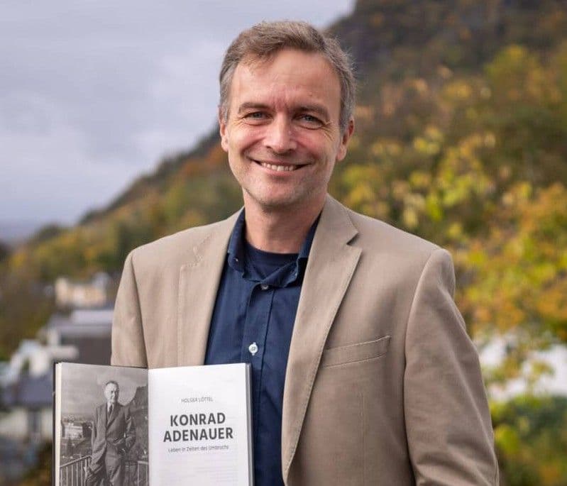 Holger Löttel in beige-farbenem Sakko hält ein aufgeschlagenes Buch in die Kamera. Auf der rechten Buchseite steht der Titel „KONRAD ADENAUER“. Im Hintergrund ist eine bewaldete Hügellandschaft unter bewölktem Himmel zu sehen.