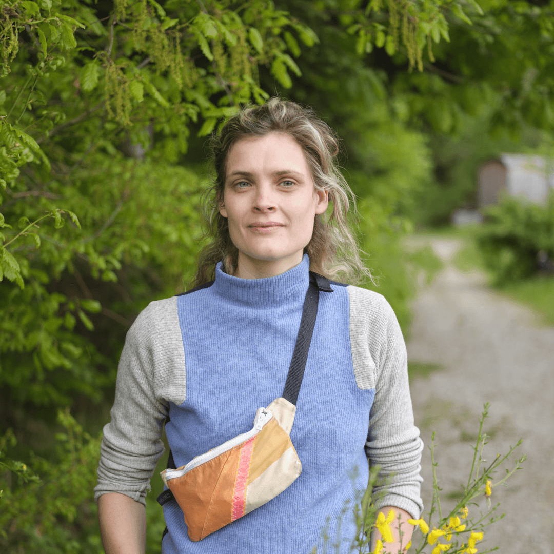 Porträtfoto einer Frau mit lockigen blonden Haaren, gekleidet in einen blau-grauen Pullover mit einer Umhängetasche in Goldtönen. Sie lächelt leicht in die Kamera. Der Hintergrund zeigt einen grünen Waldweg mit Laubbäumen im Freien.