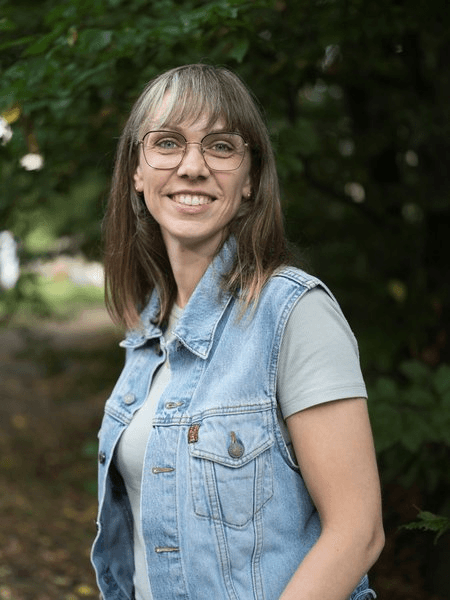 orträtfoto einer Frau mit langen braunen Haaren mit Pony und einer eckigen Brille, gekleidet in eine hellblaue Jeansweste über einem grauen T-Shirt. Sie lächelt freundlich in die Kamera. Der Hintergrund zeigt einen unscharfen Waldweg mit Bäumen.