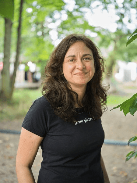 Porträtfoto einer Frau mit langen, leicht gewellten dunklen Haaren, gekleidet in ein schwarzes T-Shirt. Sie lächelt leicht in die Kamera. Der Hintergrund zeigt unscharfes grünes Laub im Freien.