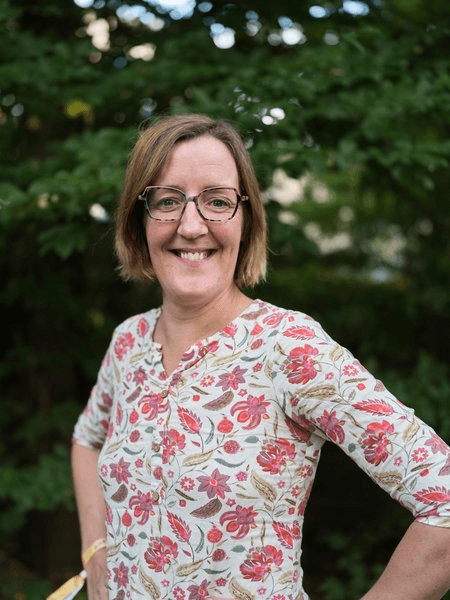 Porträtfoto einer Frau mit kurzem braunem Haar und einer Brille, gekleidet in eine weiße Bluse mit rotem Blumenmuster. Sie lächelt breit in die Kamera. Der Hintergrund zeigt unscharfes grünes Laub im Freien.