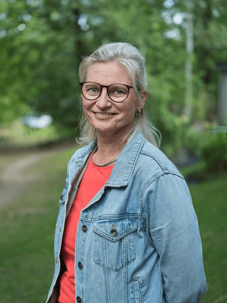 Porträtfoto einer Frau mit nach hinten gestecktem graublondem Haar und einer runden Brille, gekleidet in eine hellblaue Jeansjacke über einem roten Oberteil. Sie lächelt freundlich in die Kamera. Der Hintergrund zeigt einen unscharfen Parkweg mit Bäumen im Freien.