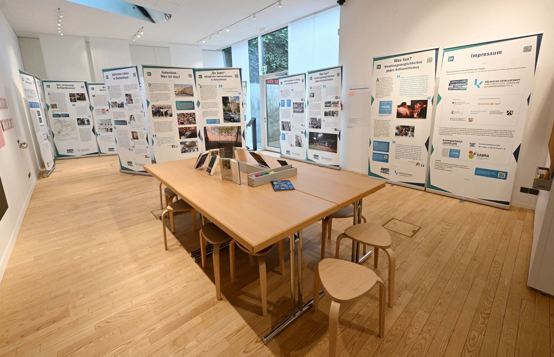 Ein heller Ausstellungsraum mit einem großen Holztisch in der Mitte, umgeben von zahlreichen Informationstafeln zum Thema Judentum und Antisemitismus.