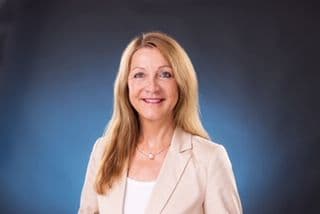 Porträtfoto einer Frau mit langen blonden Haaren und einem hellen Blazer. Sie lächelt freundlich in die Kamera. Der Hintergrund ist dunkelblau-grau und neutral gehalten.