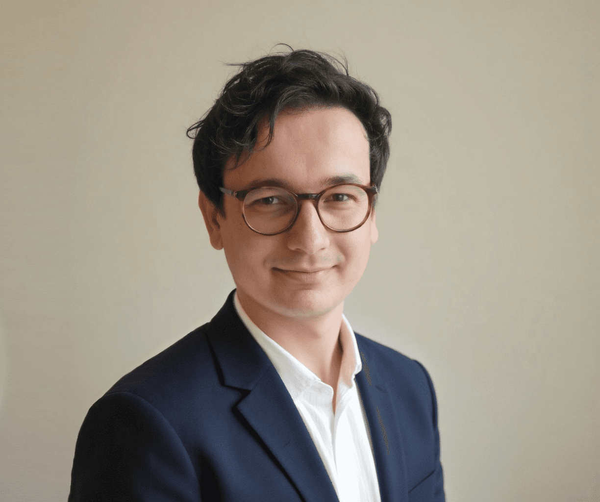 Porträtfoto eines Mannes mit dunklem lockigem Haar und einer runden Hornbrille, gekleidet in einen dunkelblauen Blazer und weißes Hemd. Er lächelt leicht in die Kamera. Der Hintergrund ist hell und neutral gehalten.
