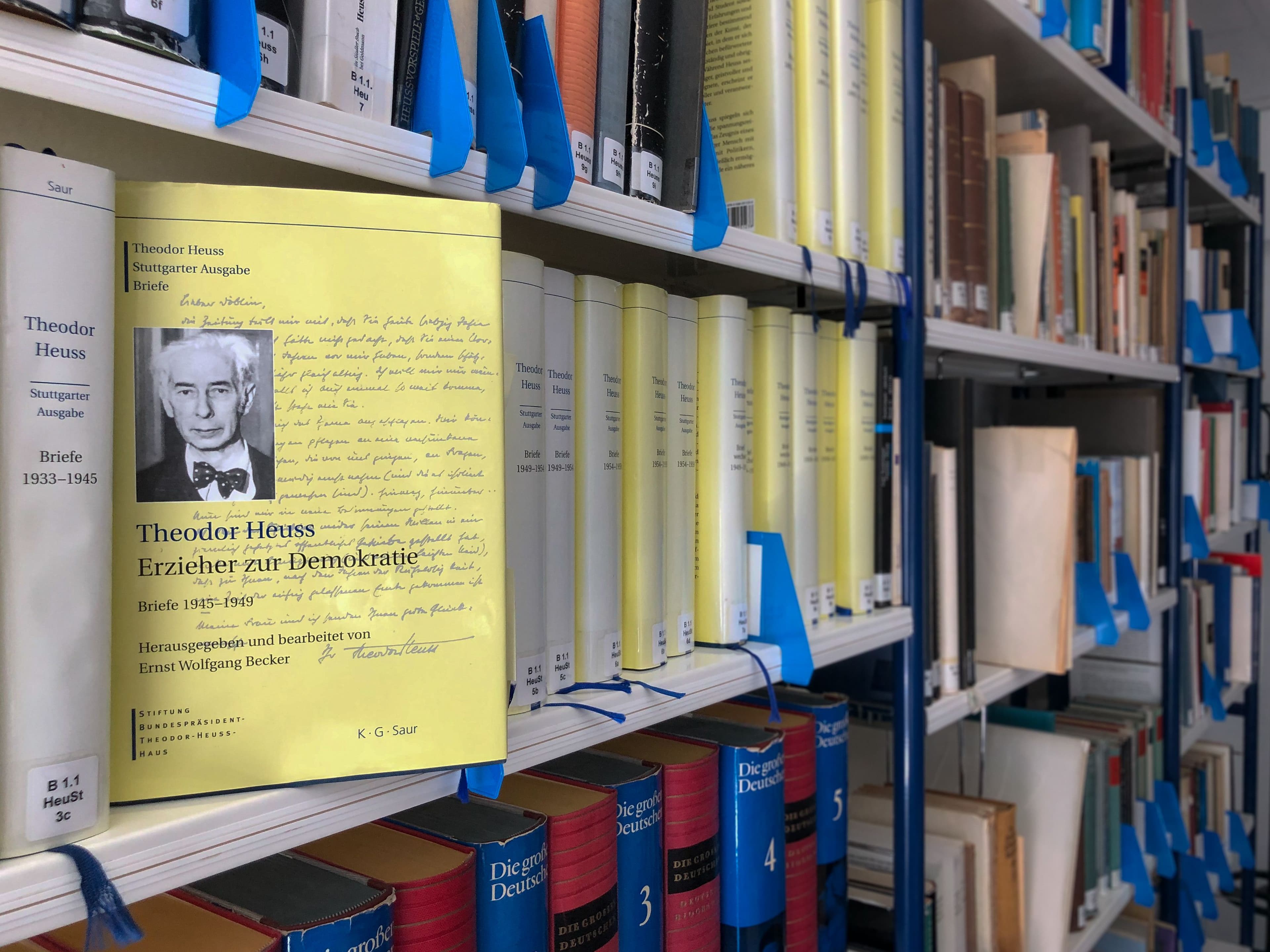 Ein Buchcover mit dem Titel „Theodor Heuss: Erzieher zur Demokratie“ steht im Vordergrund eines Bibliotheksregals, flankiert von weiteren Bänden der Stuttgarter Ausgabe.