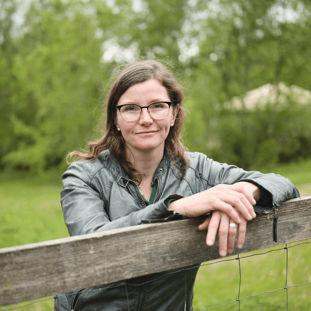 Porträtfoto einer Frau mit langen braunen Haaren und einer schwarz-transparenten Brille, gekleidet in eine grüne Jacke. Sie lehnt mit verschränkten Armen auf einem Holzzaun und lächelt leicht in die Kamera. Der Hintergrund zeigt eine grüne Wiese mit Bäumen im Freien.