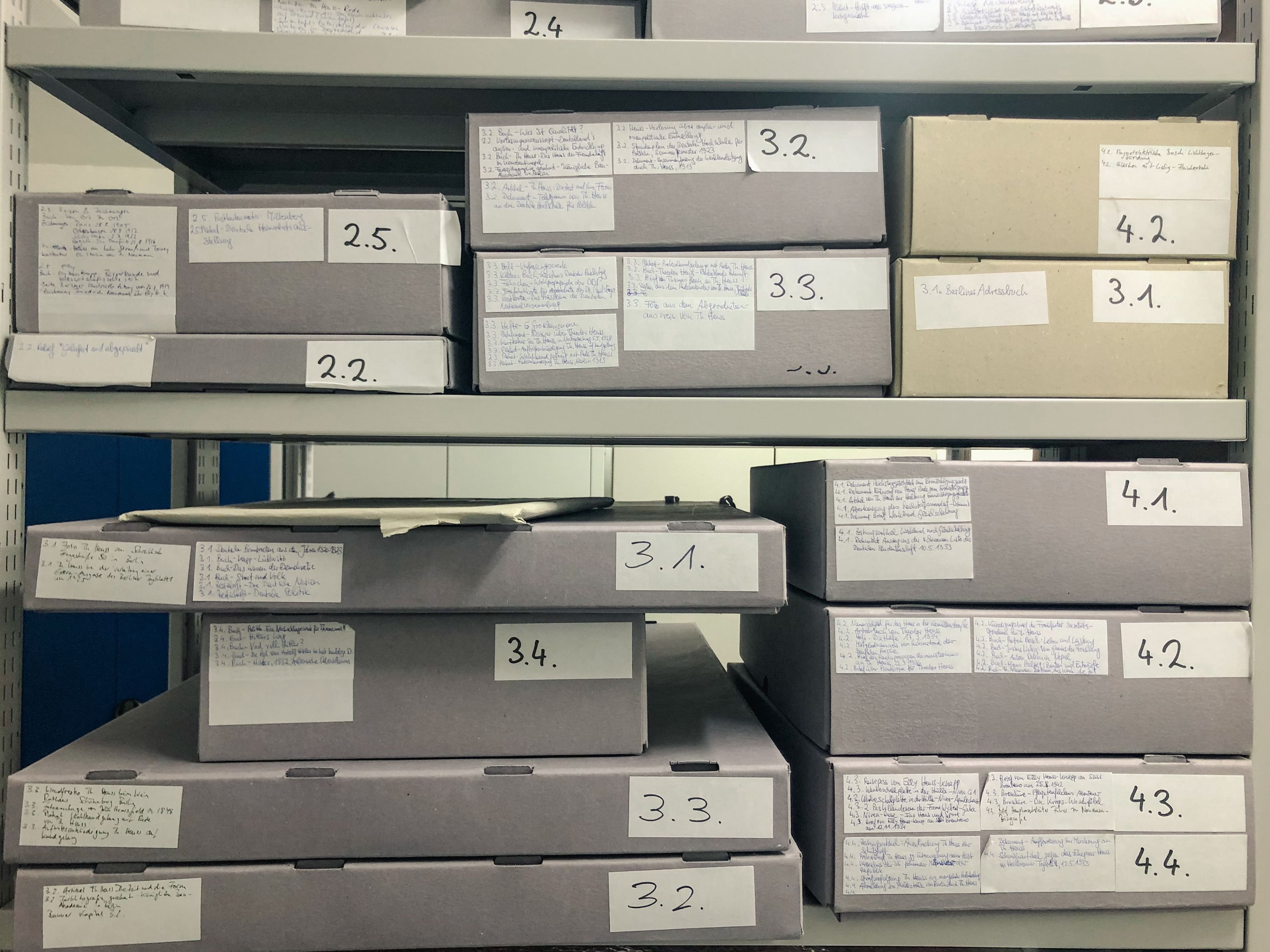 Mehrere Metallregale in einem Archiv, die mit grauen Archivkartons unterschiedlicher Größe gefüllt sind, welche handschriftliche Etiketten und Nummern tragen.