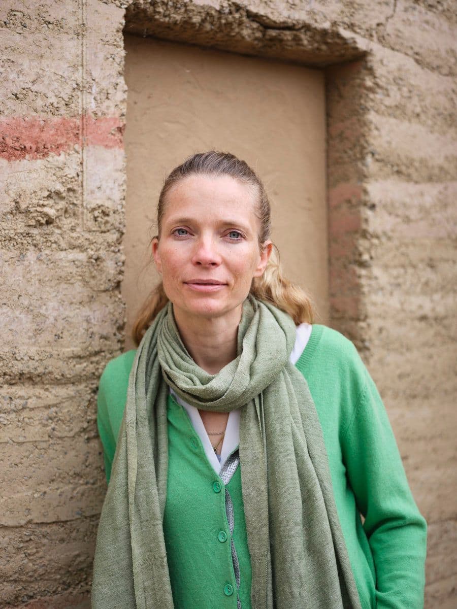 Porträtfoto einer Frau mit hochgesteckten braunen Haaren, gekleidet in eine braune Strickjacke über einer gemusterten blauen Bluse und heller Jeans. Sie steht in einem Türrahmen vor einem hellgrauen Hintergrund und lächelt leicht in die Kamera.