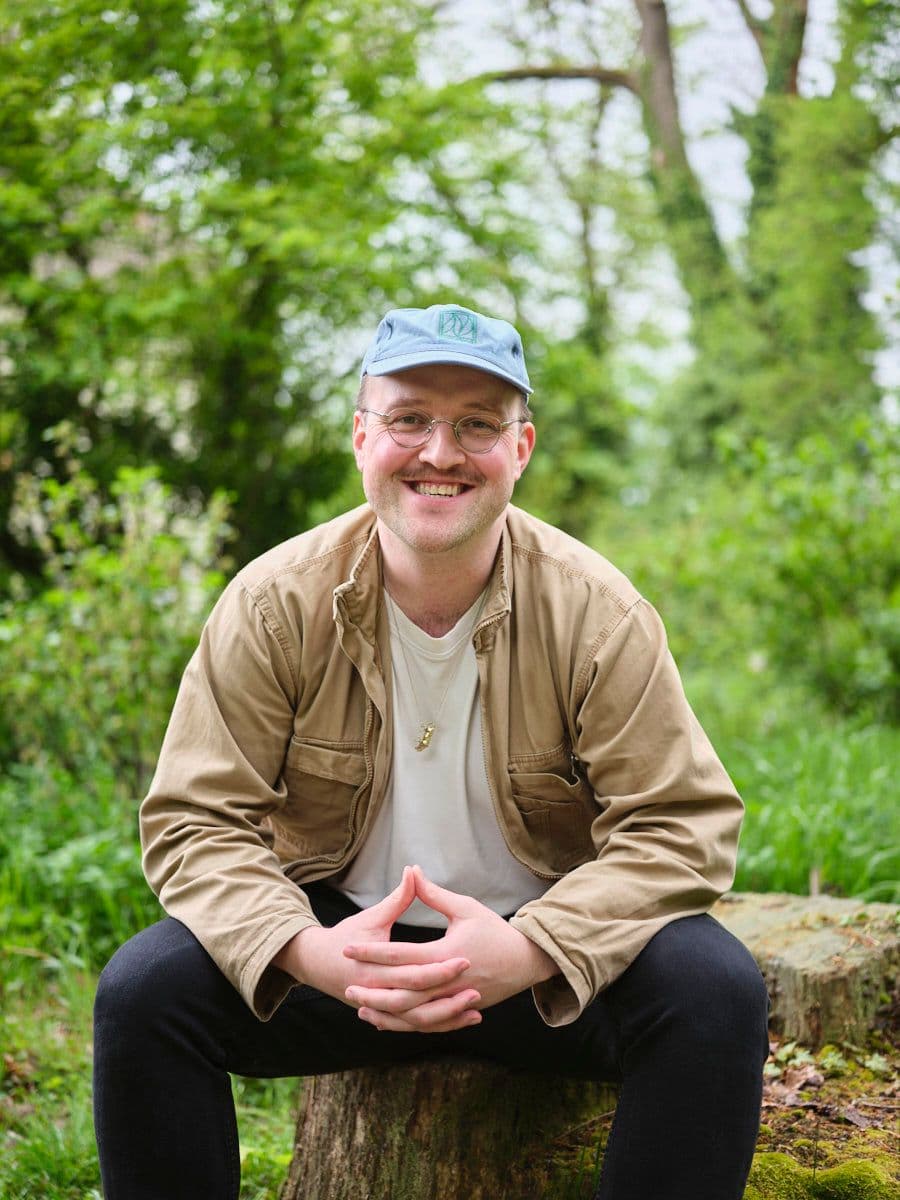 Porträtfoto eines Mannes mit einer hellblauen Cap und einer runden Brille, gekleidet in eine beige Jacke über einem weißen T-Shirt. Er sitzt auf einem Baumstumpf, hält die Hände gefaltet und lächelt freundlich in die Kamera. Der Hintergrund zeigt üppiges Grün im Freien.