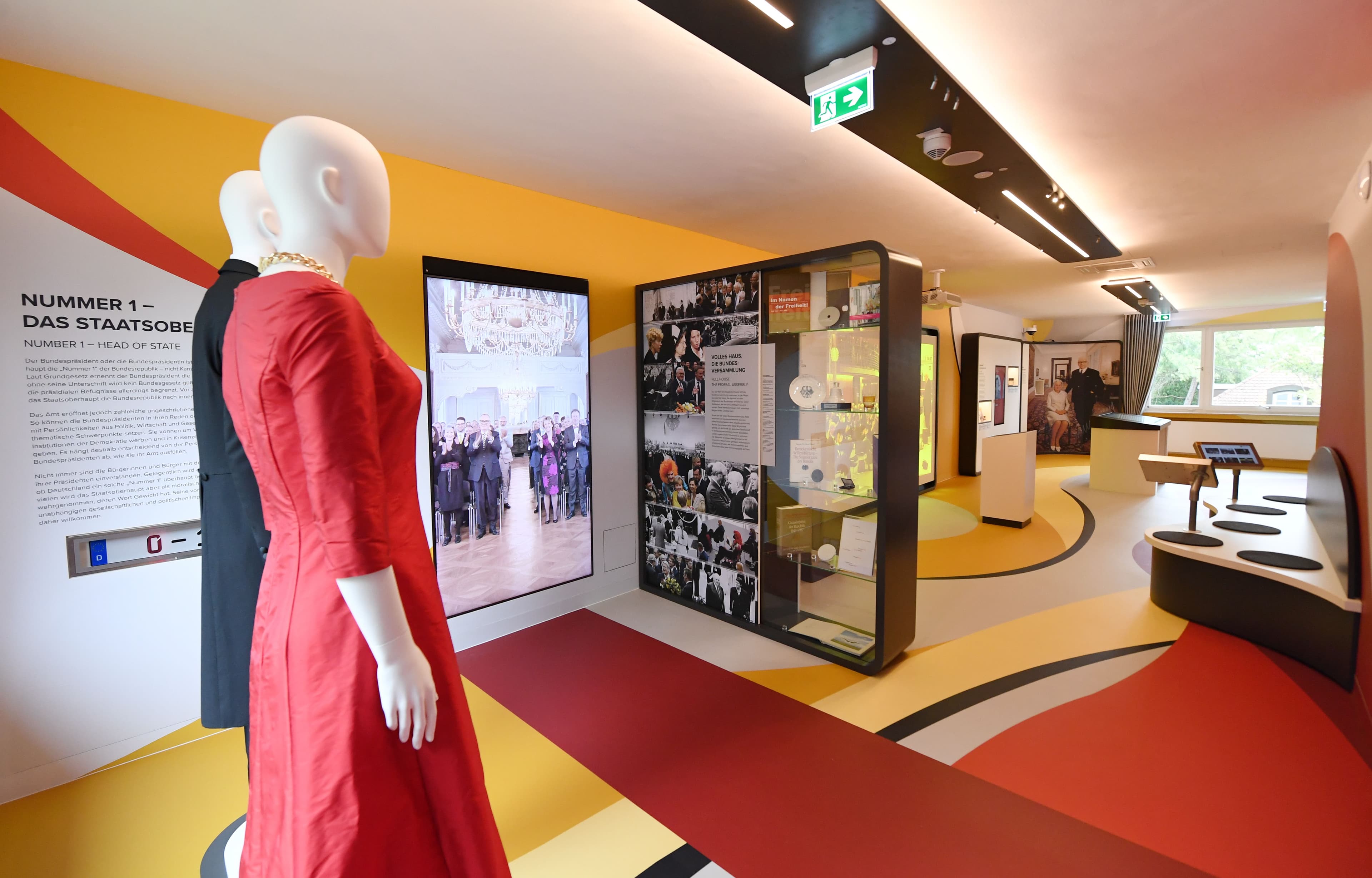 Innenansicht eines modernen Museums mit farbenfrohem Boden in den Farben Schwarz, Rot und Gold. Im Vordergrund stehen zwei Schaufensterpuppen, eine in einem roten Kleid mit goldener Halskette, eine in einem dunklen Anzug. An der Wand ist ein Ausstellungstext mit dem Titel „Nummer 1 – Das Staatsoberhaupt" zu lesen. Im Hintergrund befinden sich Vitrinen mit historischen Fotografien sowie digitale Bildschirme und interaktive Ausstellungselemente.