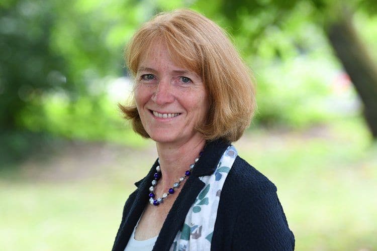 Porträtfoto einer Frau mit kurzem rotblondem Haar, einer blauen Perlenkette und einer dunklen Jacke über einem gemusterten Schal. Sie lächelt freundlich in die Kamera. Der Hintergrund zeigt unscharfes grünes Laub im Freien.