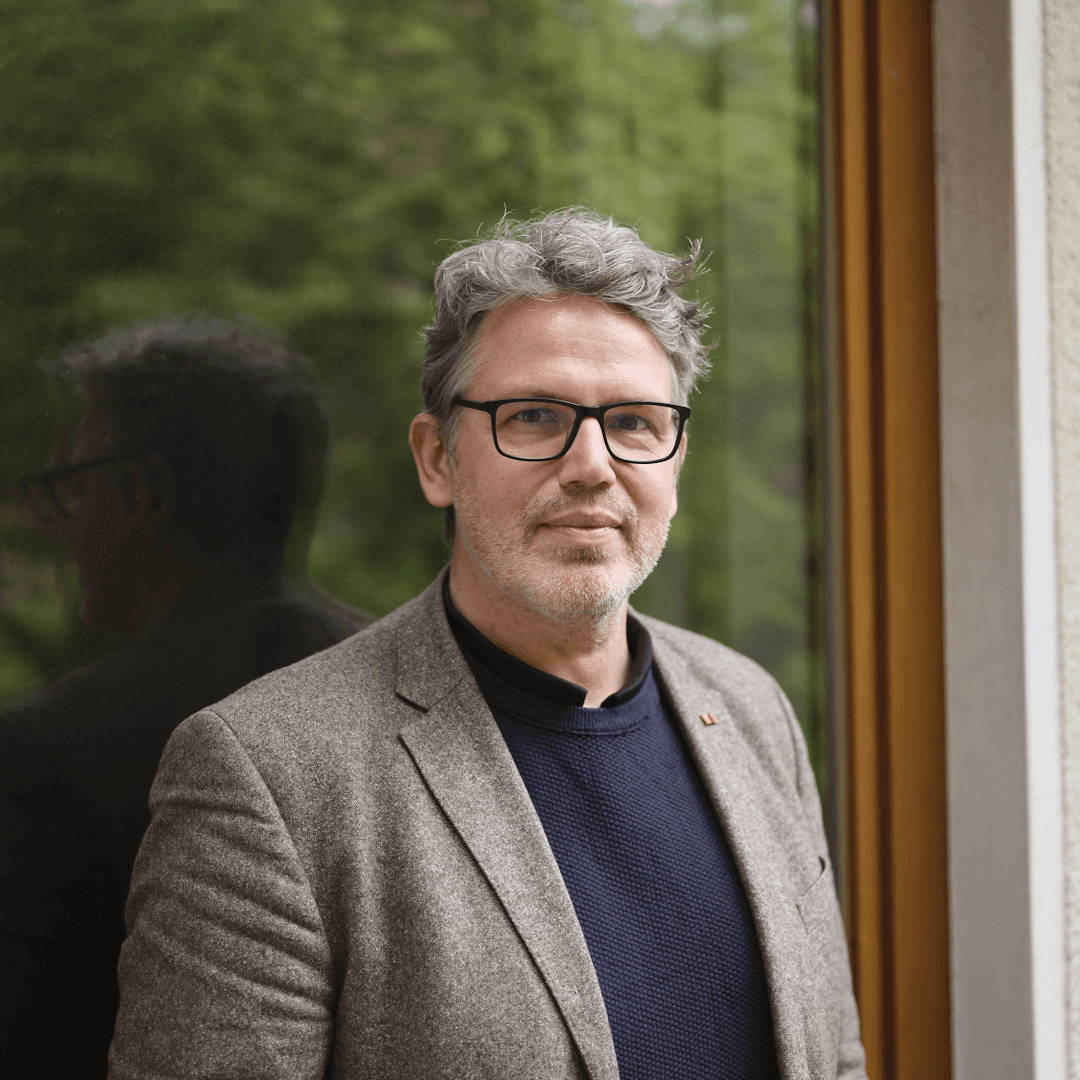 Porträtfoto eines Mannes mit grauem, leicht gewelltem Haar, Dreitagebart und einer schwarzen eckigen Brille, gekleidet in einen beigen Blazer über einem dunklen Pullover. Er lehnt an einem Fenster, durch das grünes Laub zu sehen ist, und blickt ruhig in die Kamera.