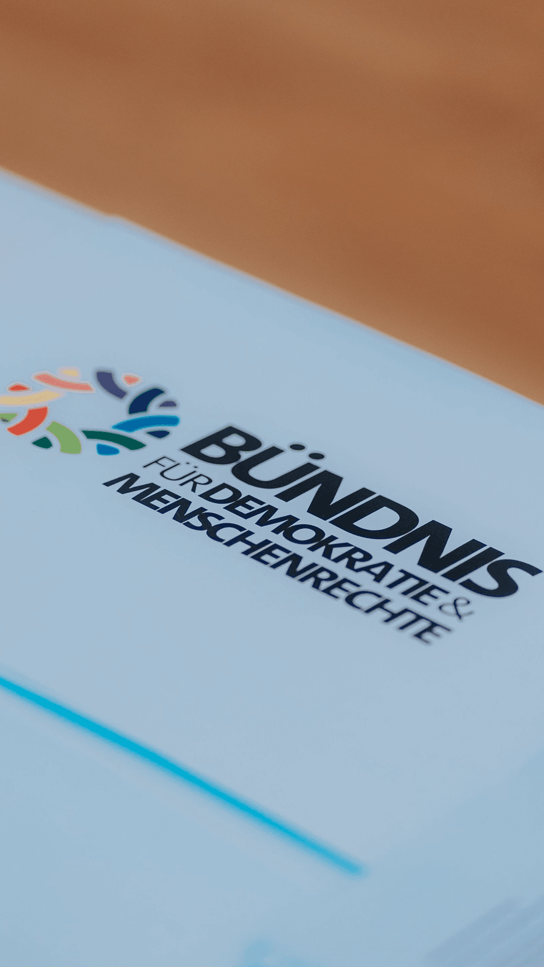 Nahaufnahme einer hellblauen Mappe, auf der das Logo und der Schriftzug „Bündnis für Demokratie & Menschenrechte" abgebildet sind. Das Logo besteht aus mehrfarbigen, stilisierten Figuren in einem Kreis. Die Mappe liegt leicht schräg auf einer Holzoberfläche.