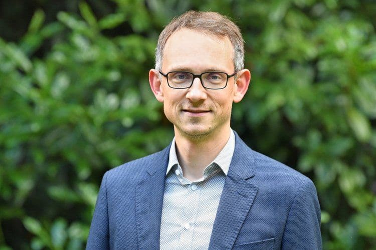 Porträtfoto eines Mannes mit kurzem hellbraunem Haar und einer schwarzen Brille, gekleidet in einen blauen Blazer und ein gemustertes Hemd. Er lächelt leicht in die Kamera. Der Hintergrund zeigt unscharfes grünes Laub im Freien.
