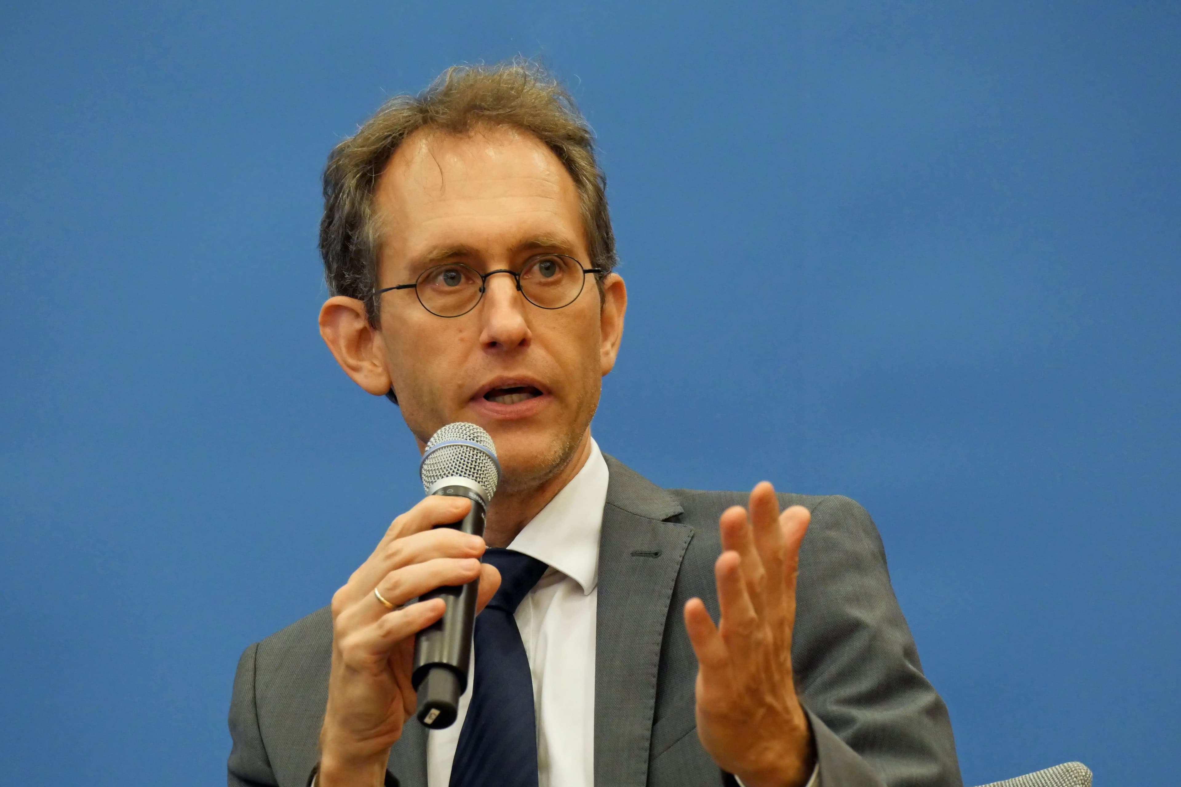 Frieder Günther mit Brille, kurzem braunem Haar und grauem Anzug mit dunkler Krawatte spricht in ein Mikrofon. Er hält das Mikrofon in der rechten Hand und unterstreicht seine Aussage mit einer Geste der linken Hand. Der Hintergrund ist einflächig blau.