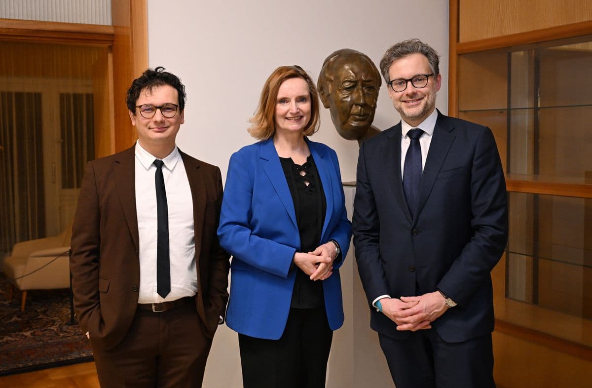 drei Personen, links ein Mann in braunem Anzug und Krawatte, in der Mitte eine Frau in blauem Blazer, rechts ein Mann mit dunklem Anzug und Krawatte