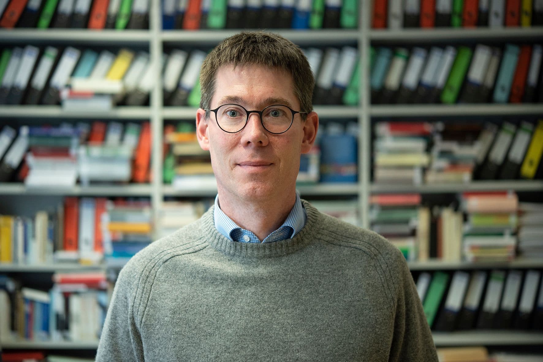 Porträt von Christopher Dowe mit Brille und kurzem braunem Haar vor einem gut gefüllten Akten- und Bücherregal. Er trägt einen grauen Strickpullover über einem hellblauen Hemd.