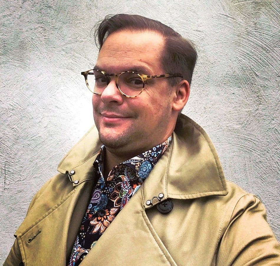 ein lächelnder Herr mit Brille, buntem Hemd und Trenchcoat